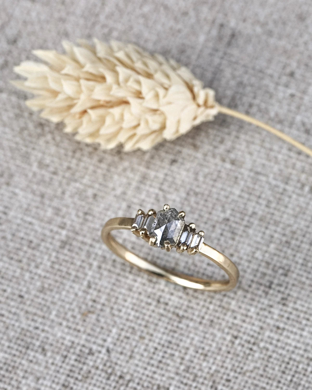 A gold ring with a cluster of baguette-cut gray gemstones, displayed on a textured gray linen background next to a dried wheat stem.