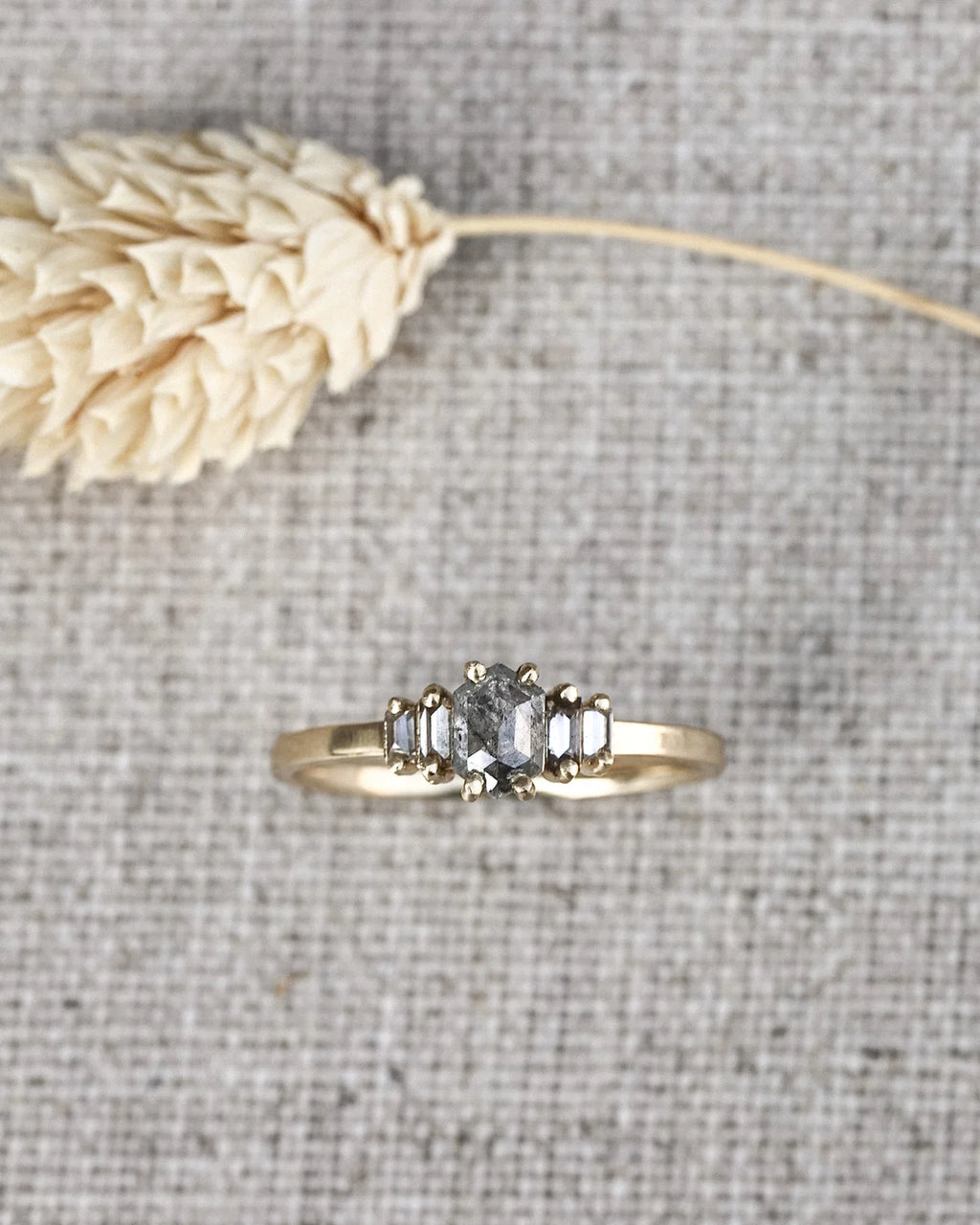 A yellow gold ring with a central round-cut clear gemstone and two baguette-cut side stones, displayed on a textured gray linen background with a dried beige flower stem.
