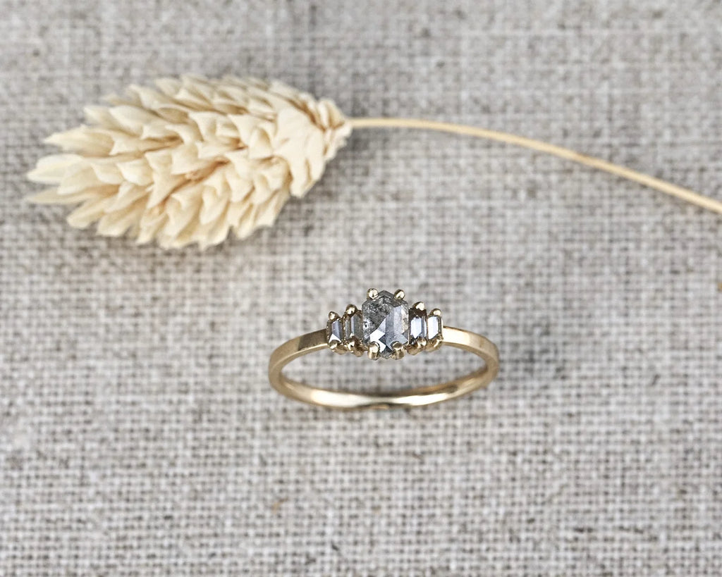 A gold three-stone ring with a central oval clear gemstone and two marquise-cut side stones, displayed on a textured linen surface next to a dried flower stem.