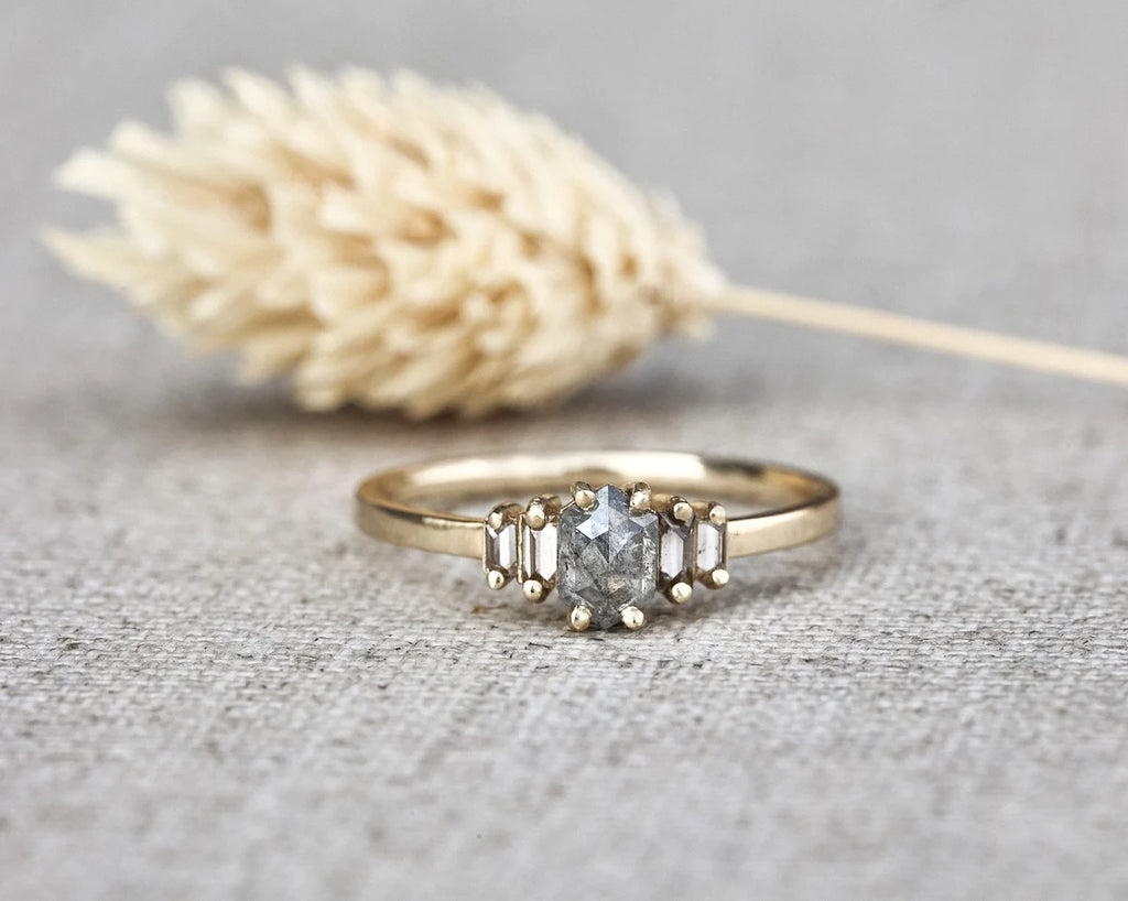 A gold ring with a central round clear gemstone and two baguette-cut side stones, displayed on a textured gray fabric surface with a blurred dried grass stem in the background.