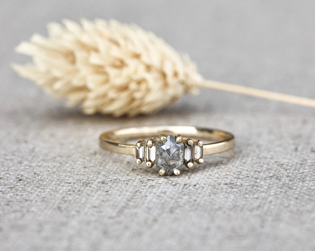 A gold ring with a central round clear gemstone and two baguette-cut side stones, displayed on a textured gray fabric surface with a blurred dried grass stem in the background.