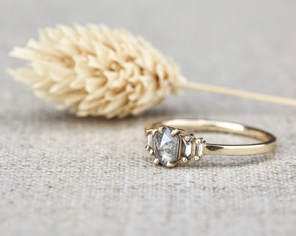 A gold ring with a central clear gemstone and small side stones, displayed on a textured beige fabric surface with a dried beige flower in the background.