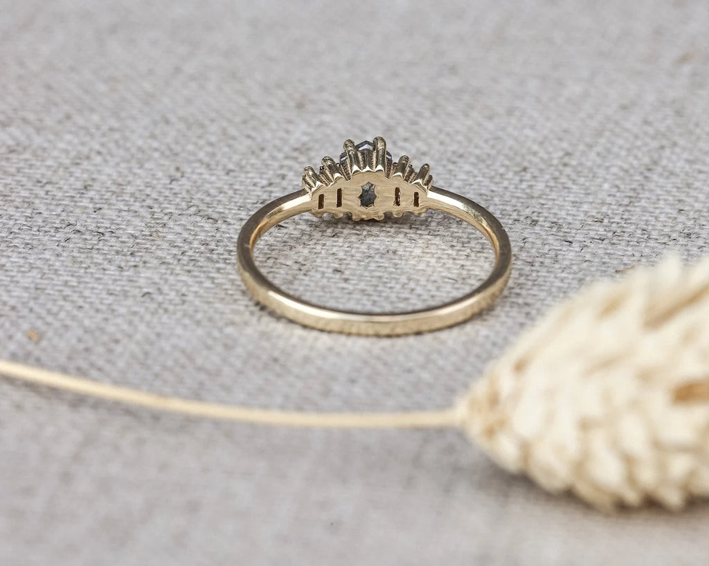 A gold ring with a crown-like top and a small dark center stone, resting on a textured beige fabric surface with a dried flower in the foreground.