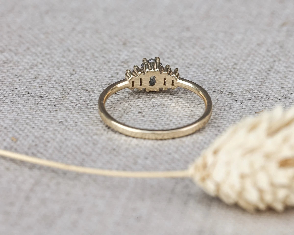 A gold ring with a crown-like top and a small dark center stone, resting on a textured beige fabric surface with a dried flower in the foreground.