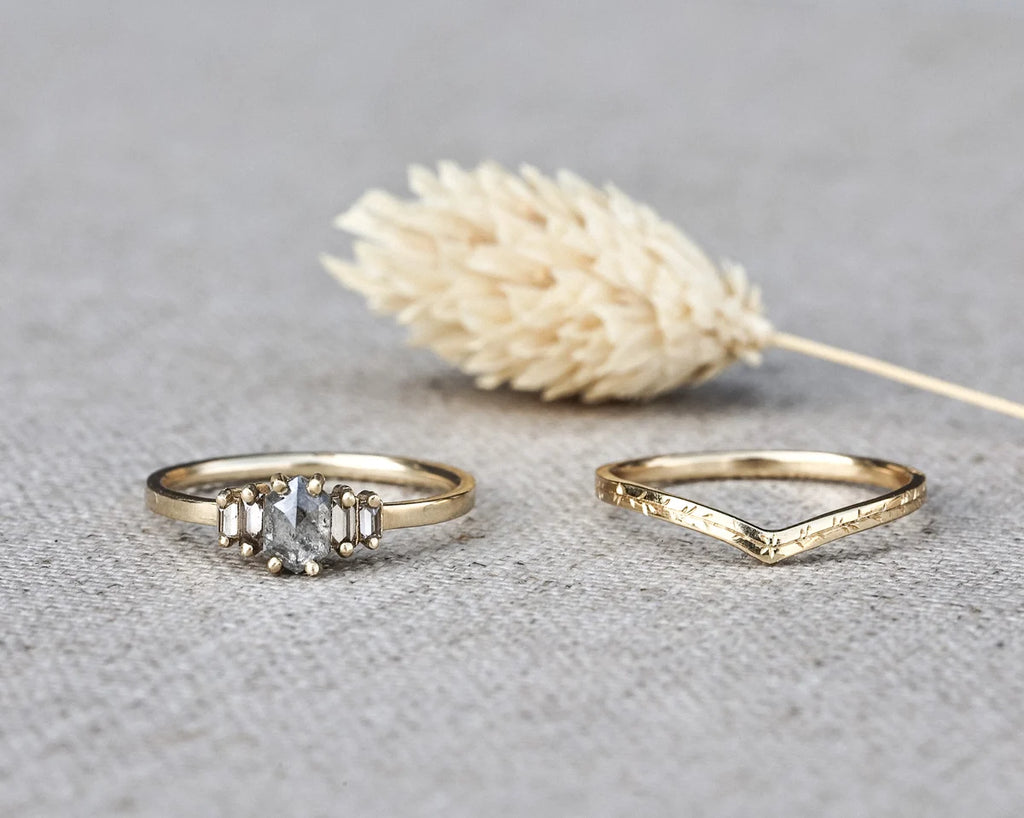 Two gold rings displayed on a textured gray surface with a dried beige flower in the background. One ring features a clear baguette-cut center stone with smaller side stones, and the other has a textured, hammered geometric band.