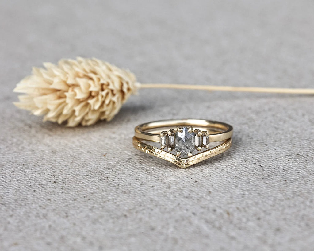 A gold ring with a central clear rectangular gemstone and two smaller side stones, displayed on a textured gray surface next to a dried beige flower.