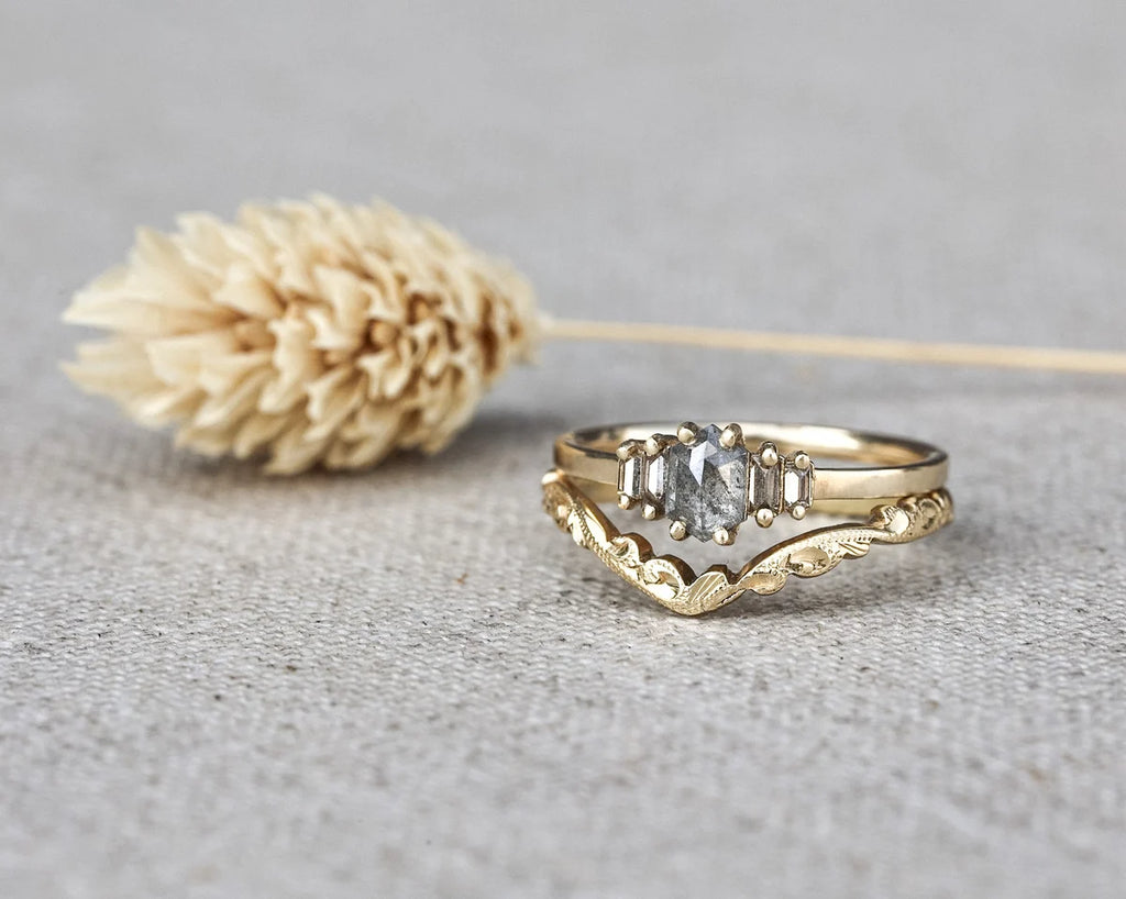 A gold ring with a central clear gemstone and decorative scrollwork, displayed on a textured gray surface next to a dried beige flower.