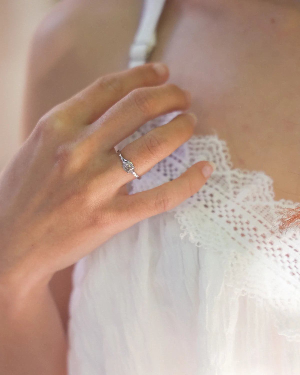 A close-up lifestyle shot of a woman wearing a delicate silver ring with a small clear gemstone on her finger, resting her hand on her white lace-trimmed top.