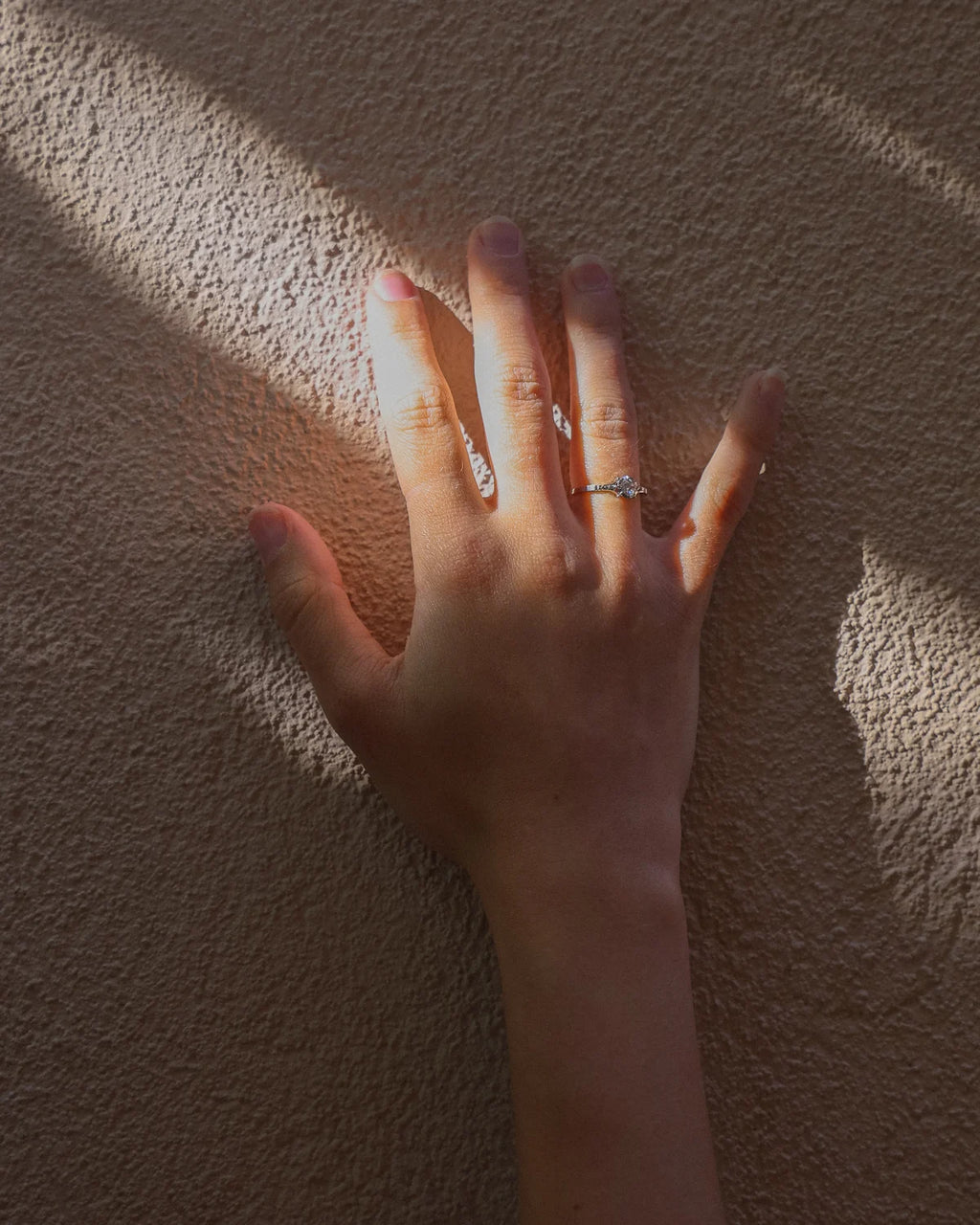 A close-up of a person's hand resting against a textured beige wall, wearing a delicate silver ring on one finger.