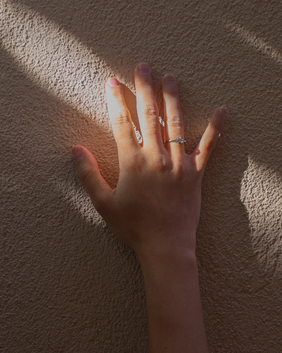 A close-up of a person's hand resting against a textured beige wall, wearing a delicate silver ring on one finger.