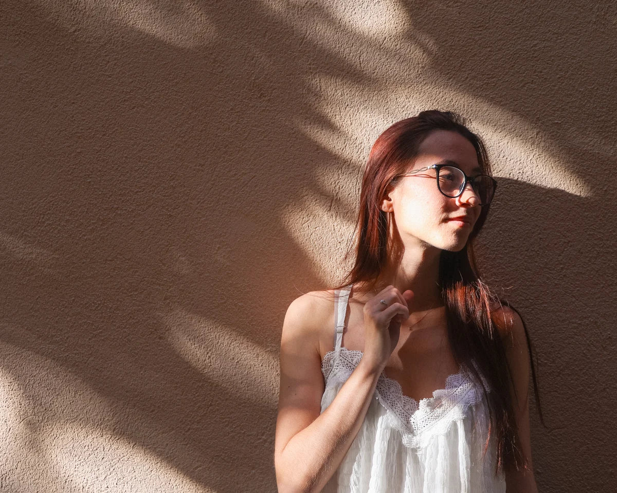 A woman with long reddish-brown hair and glasses wearing a white lace-trim camisole top, standing against a textured beige wall with dappled sunlight and shadows.