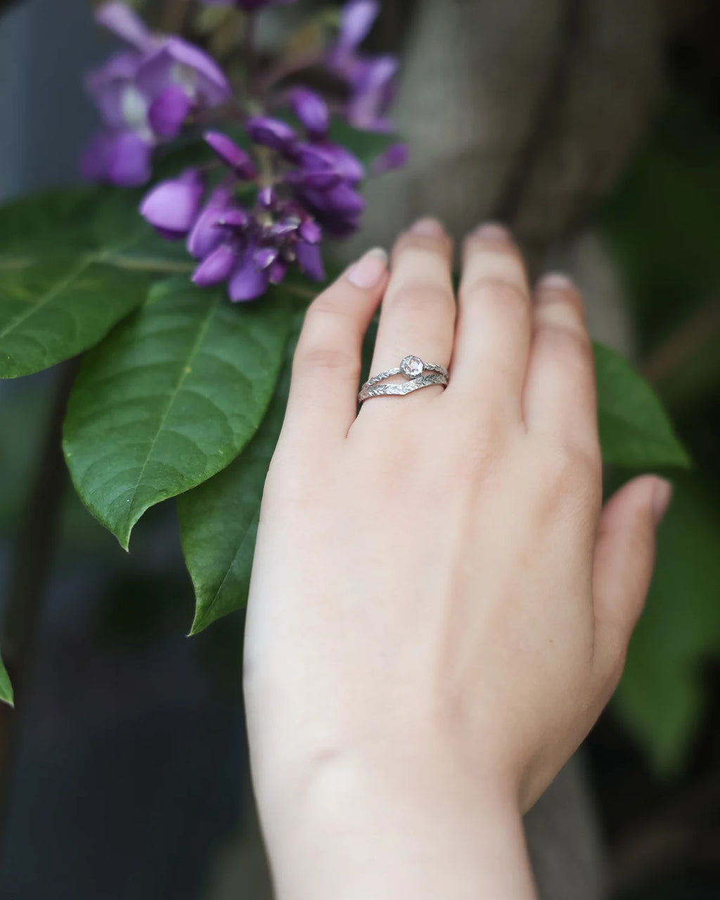 A close-up of a person's hand wearing a platinum band ring with a round clear gemstone, set against a soft-focus background of green leaves and purple flowers.