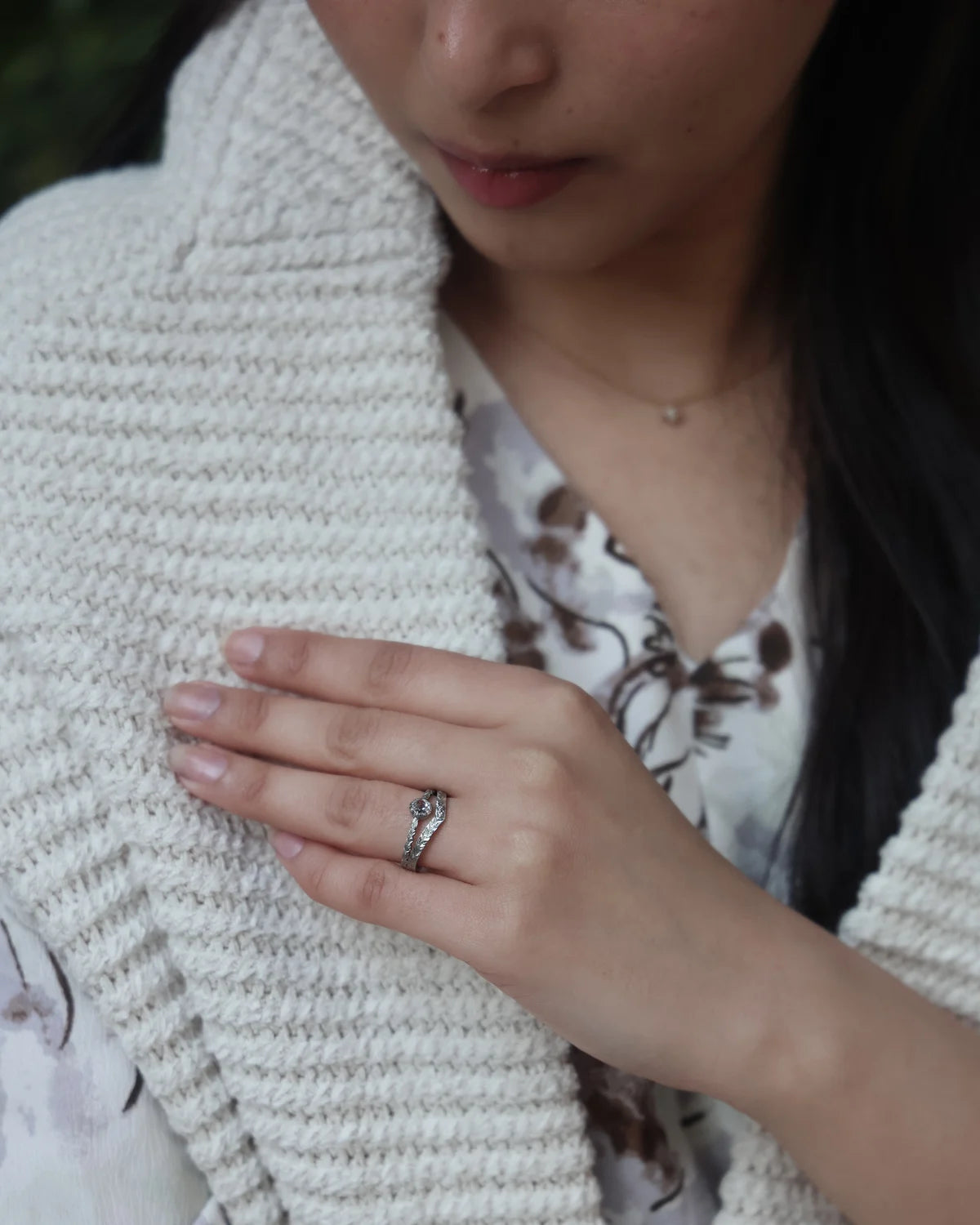 A close-up of a person wearing a platinum ring with small clear stones, shown on their hand against a textured cream knit cardigan and floral blouse.
