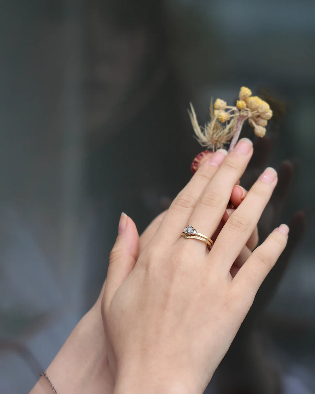 A delicate gold ring with a small clear gemstone is worn on a person's ring finger, shown against a soft, blurred dark gray background with dried yellow flowers resting on the hand.