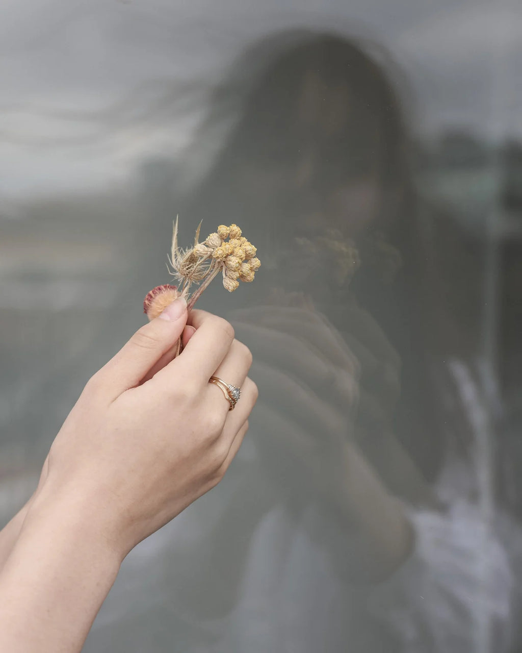 A close-up of a hand holding a small dried flower sprig in front of a reflective, smoky gray surface with a blurred reflection of a person.