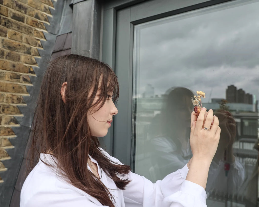 A woman with long brown hair wearing a white shirt stands in profile next to a large window, holding a small dried flower in her hand. The scene is set against a brick wall and the window reflection shows her silhouette against an overcast sky.