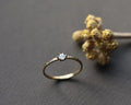 A delicate gold ring with a single round clear gemstone in a prong setting, displayed on a dark gray surface next to dried yellow flowers.