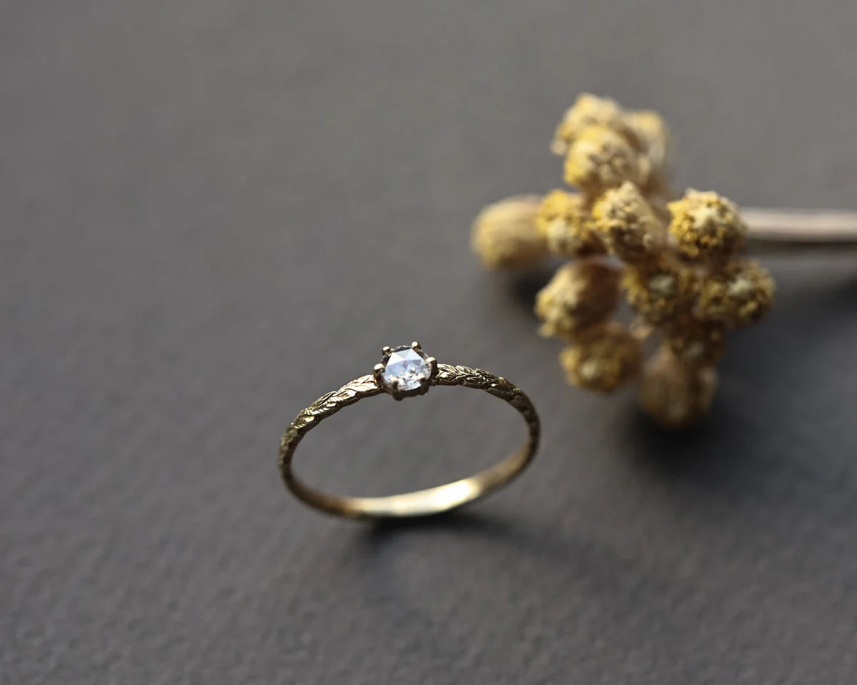 A delicate gold ring with a single round clear gemstone in a prong setting, displayed on a dark gray surface next to dried yellow flowers.