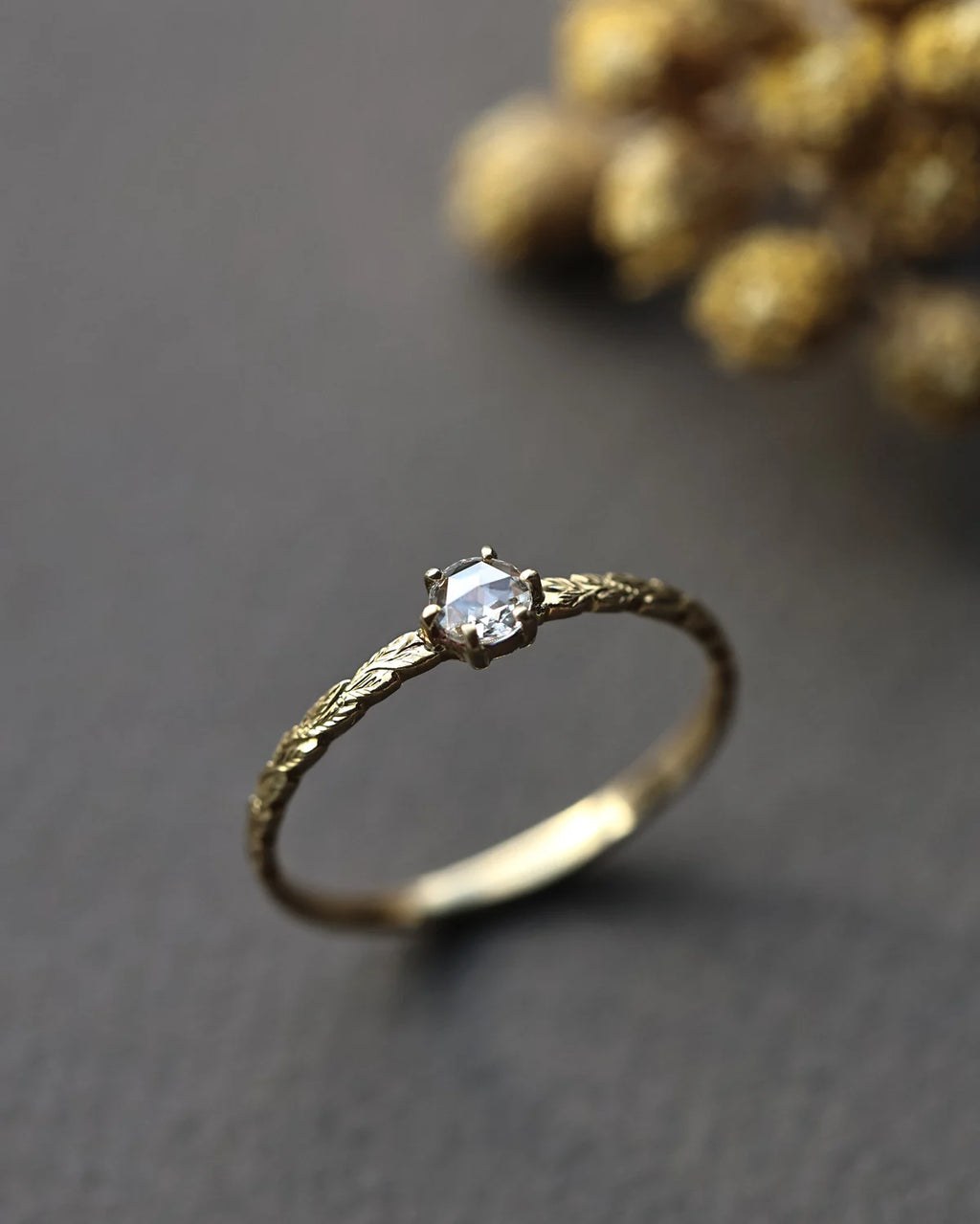 A delicate gold ring with a textured band and a single round clear gemstone, photographed on a dark gray surface with a blurred gold decorative element in the background.