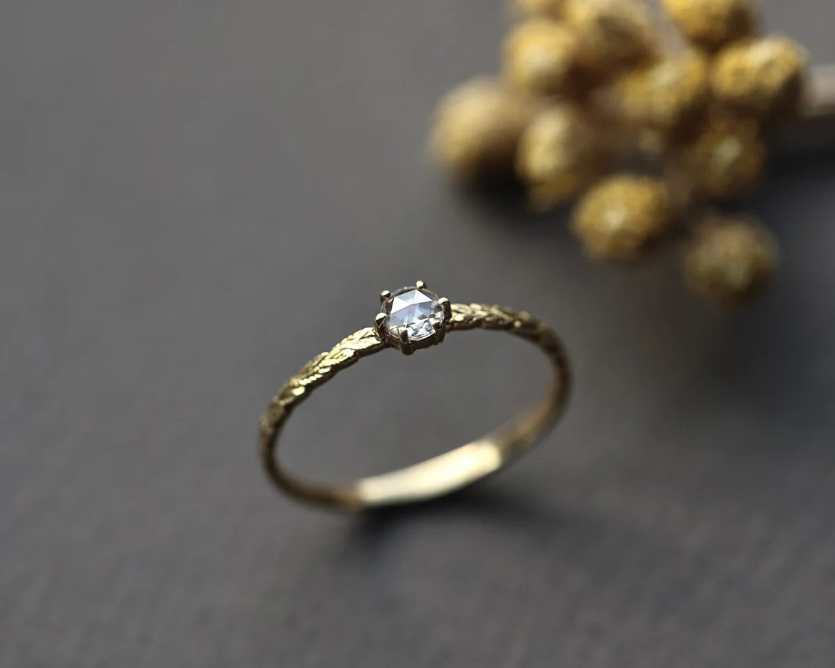 A gold ring with a single clear gemstone, photographed on a dark gray surface with a blurred cluster of small gold beads in the background.