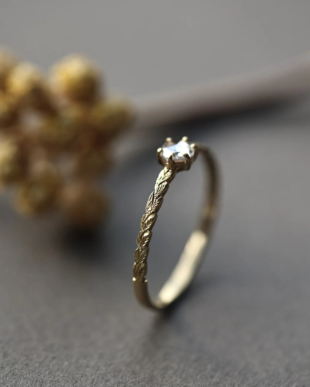 A gold ring with a textured, twisted band and a single clear gemstone in a prong setting, photographed on a dark gray surface with a blurred decorative background.