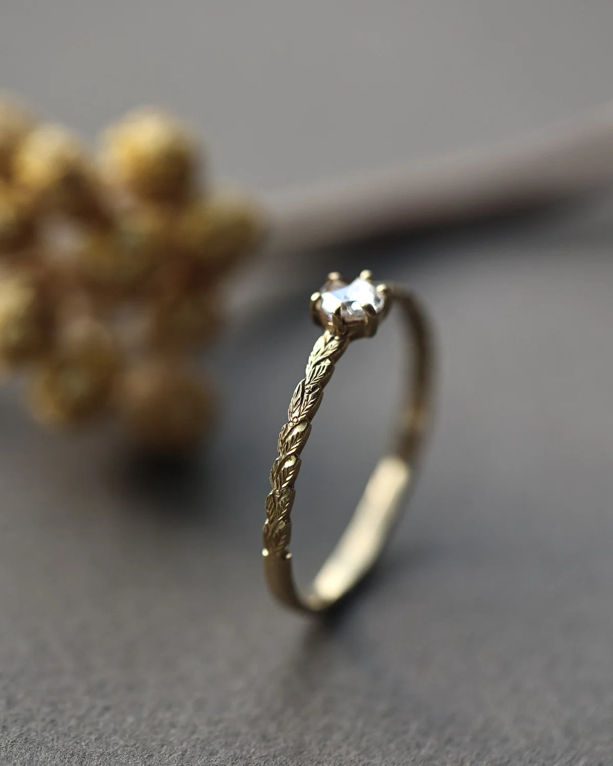A gold ring with a textured, twisted band and a single clear gemstone in a prong setting, photographed on a dark gray surface with a blurred decorative background.