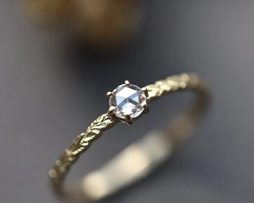 A delicate gold ring featuring a round-cut clear gemstone in a prong setting, with a textured twisted band, shown in a close-up macro shot against a dark gray background.