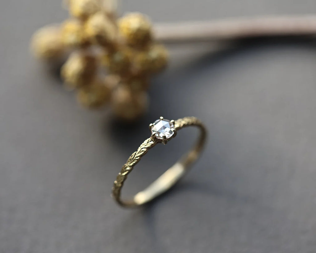 A delicate gold ring with a single clear gemstone in a prong setting, resting on a dark gray surface with a blurred dried floral background.