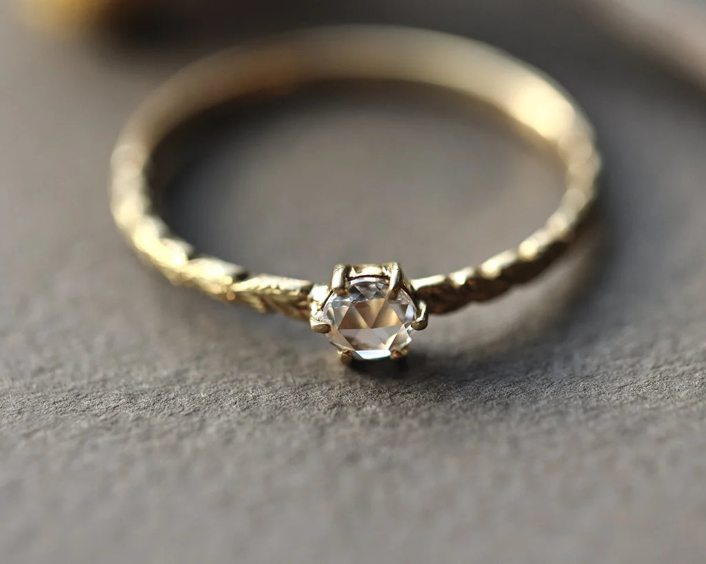 A delicate gold ring with a textured band and a central clear faceted gemstone in a prong setting, resting on a textured gray surface.