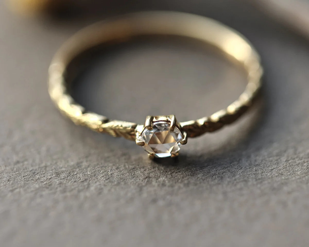 A delicate gold ring with a textured band and a central clear faceted gemstone in a prong setting, resting on a textured gray surface.