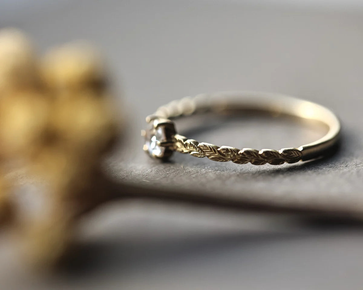 A delicate gold ring with a textured twisted band and a small clear gemstone, resting on a dark gray surface with a blurred gold decorative object in the foreground.