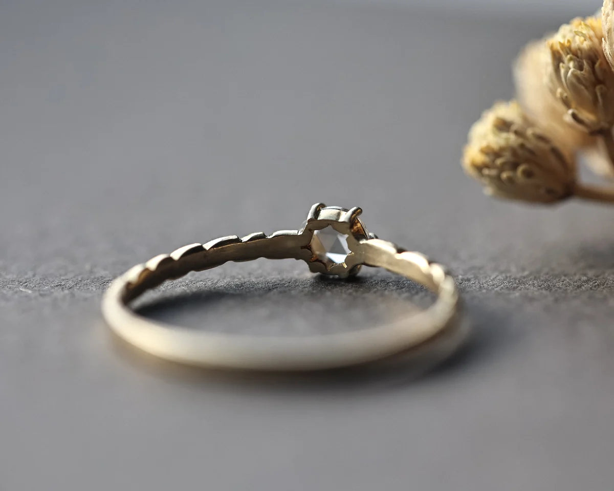 A delicate gold ring with a textured band and a small central geometric setting, photographed on a dark gray surface with dried flowers in the background.
