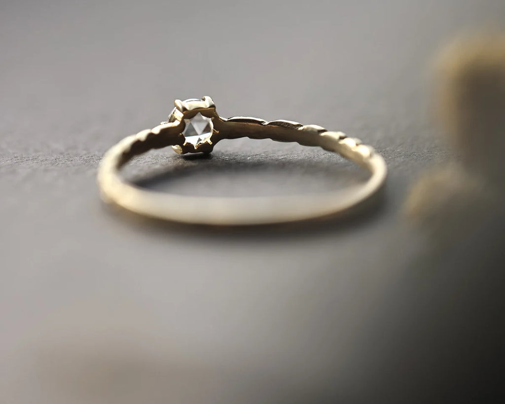 A delicate gold ring with a textured band and a small clear gemstone in a prong setting, resting on a dark gray surface.