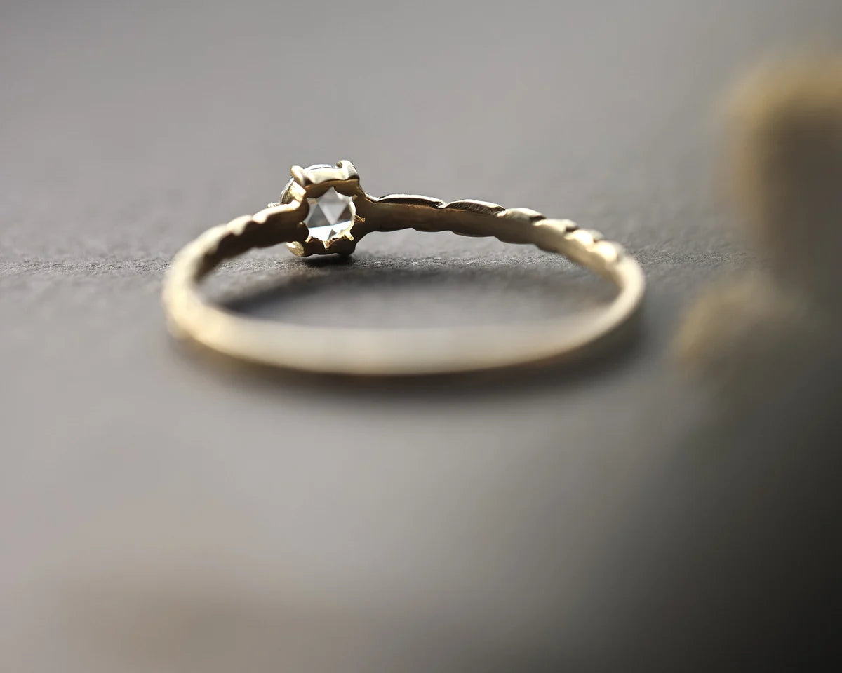 A delicate gold ring with a textured band and a small clear gemstone in a prong setting, resting on a dark gray surface.