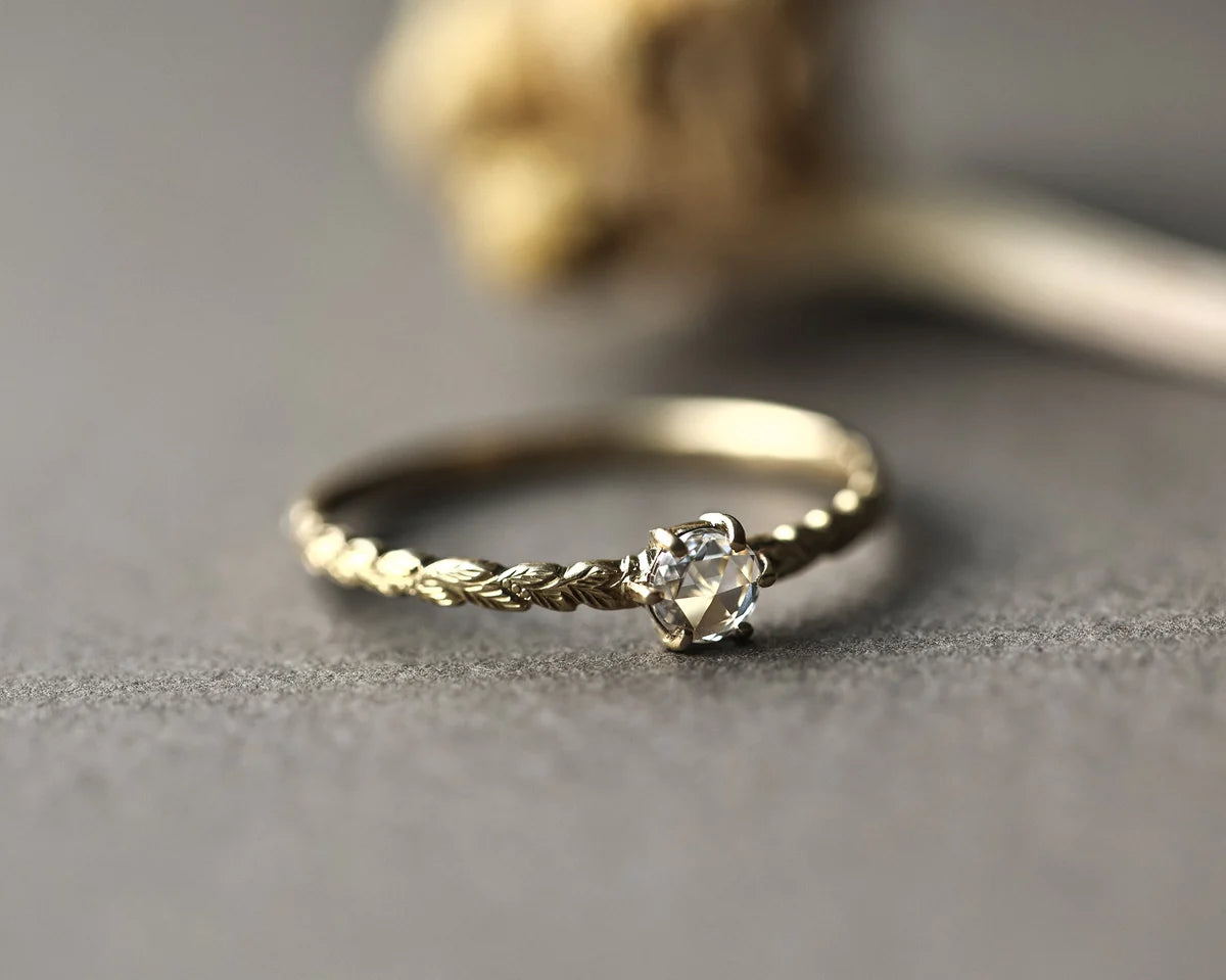 A delicate gold ring with a textured twisted band and a clear flower-shaped gemstone, resting on a gray textured surface.