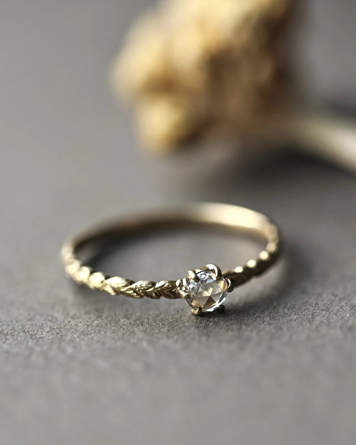 A delicate gold ring with a twisted band and a small clear heart-shaped gemstone, resting on a textured gray surface with a blurred dried flower in the background.
