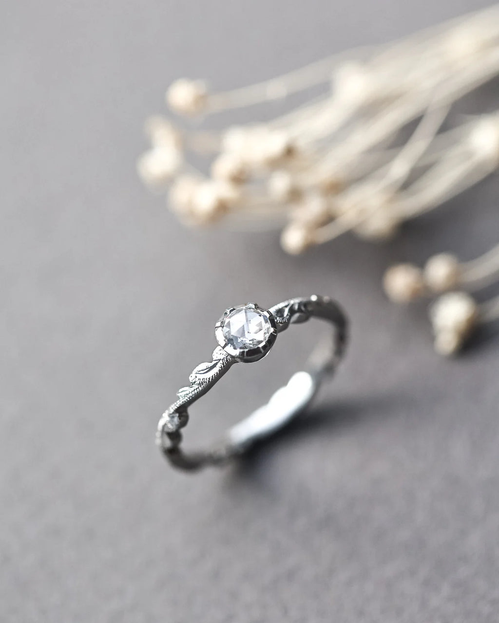 A delicate silver ring with a single round clear gemstone and ornate textured detailing on the band, photographed on a soft gray surface with blurred dried flowers in the background.
