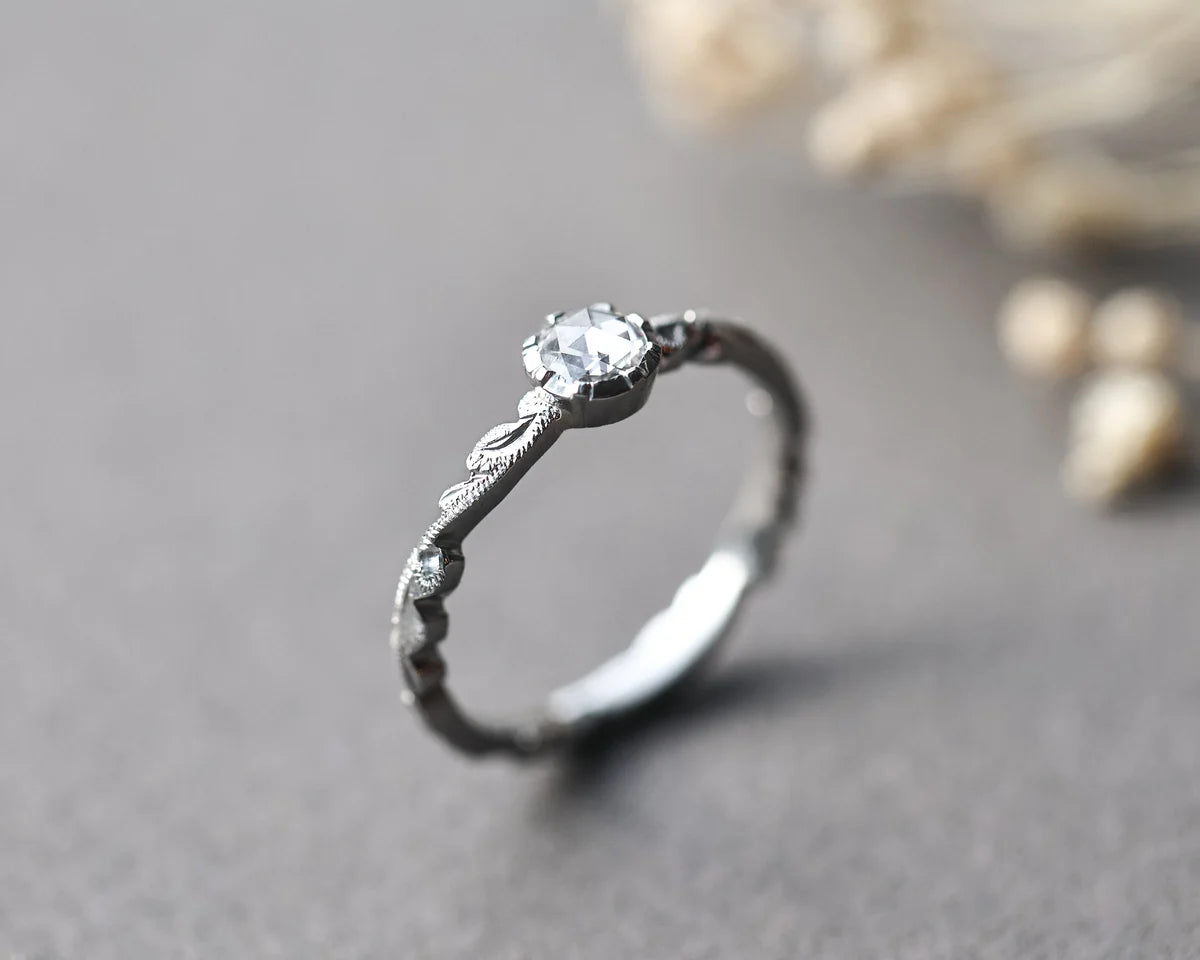A silver ring with a delicate leaf-patterned band and a round clear gemstone, photographed on a soft gray surface with blurred dried flowers in the background.