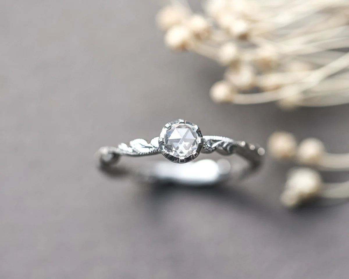 A silver ring with a round clear gemstone and a delicate leaf-like filigree band, resting on a dark gray surface with blurred dried flowers in the background.