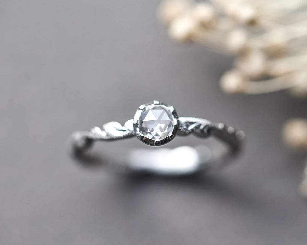 A delicate silver ring with a faceted clear gemstone and ornate twisted band, photographed on a dark gray surface with soft blurred dried flowers in the background.