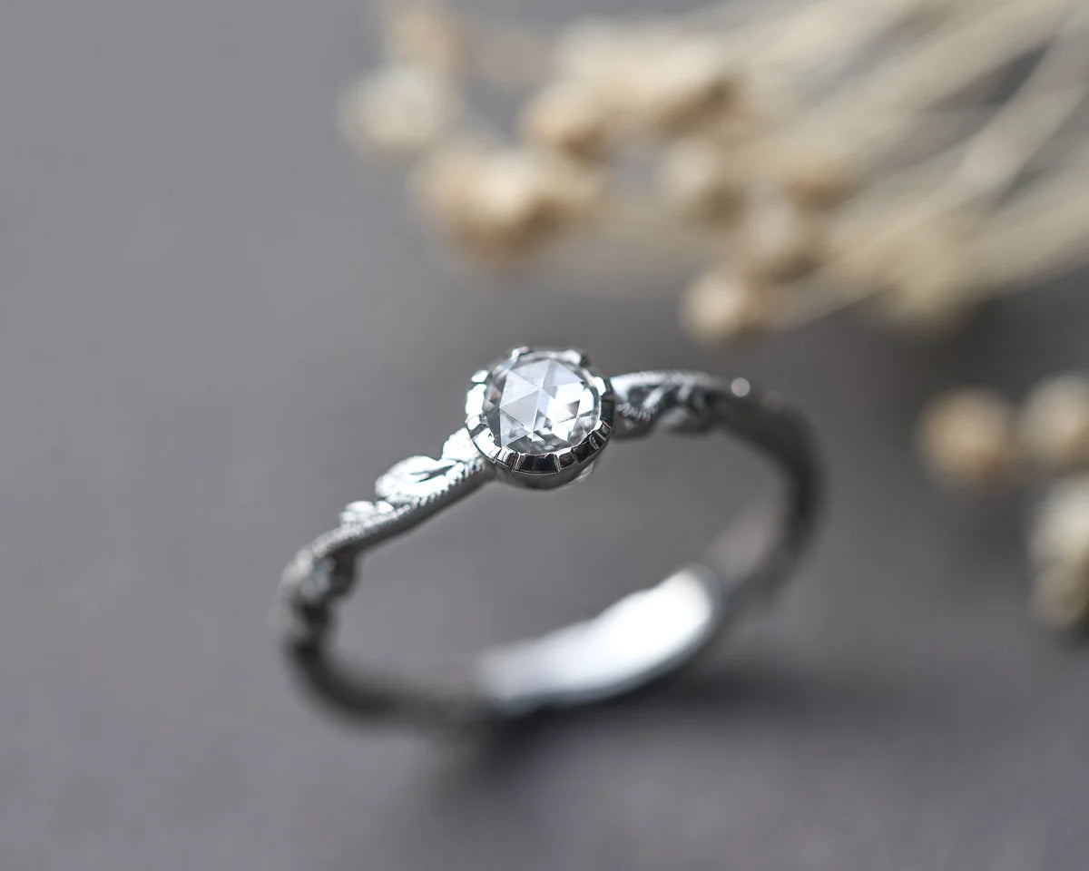 A silver-toned ring with a delicate floral or leaf-patterned band and a round clear gemstone, photographed close up on a dark gray surface with soft blurred dried flowers in the background.