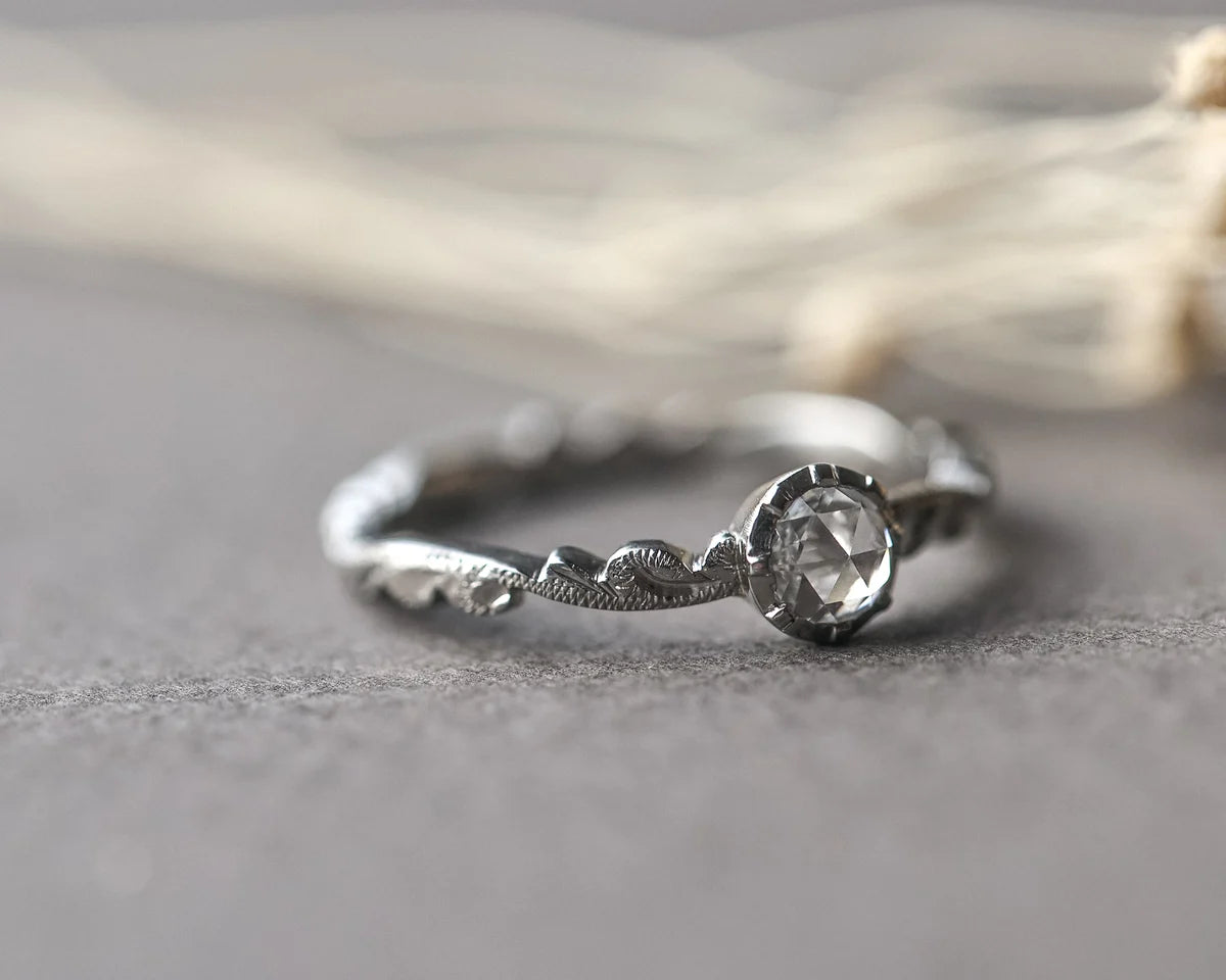 A silver ring with a textured, organic band and a faceted clear gemstone in a bezel setting, resting on a textured gray surface with a soft-focus background.