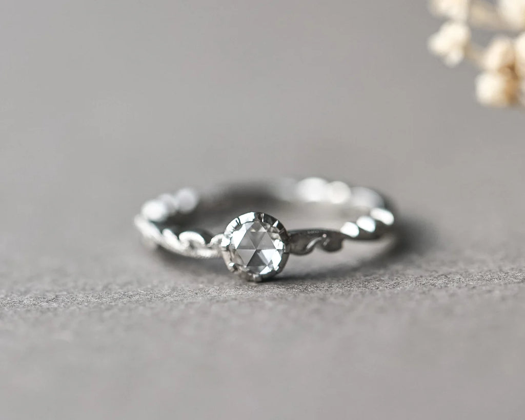 A silver-toned ring with a twisted band and a central clear faceted gemstone, photographed on a soft gray surface with a blurred floral prop in the upper right.