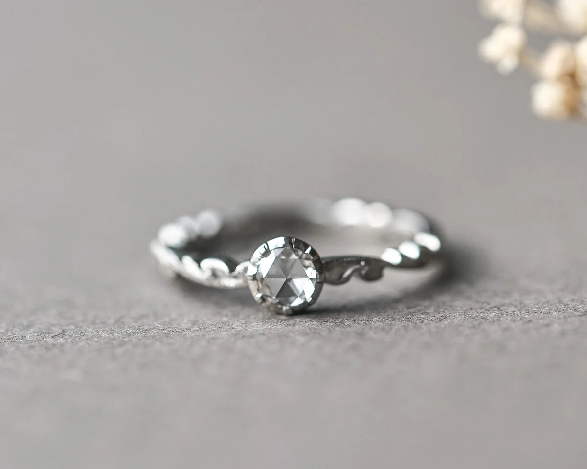 A silver-toned ring with a twisted band and a central clear faceted gemstone, photographed on a soft gray surface with a blurred floral prop in the upper right.