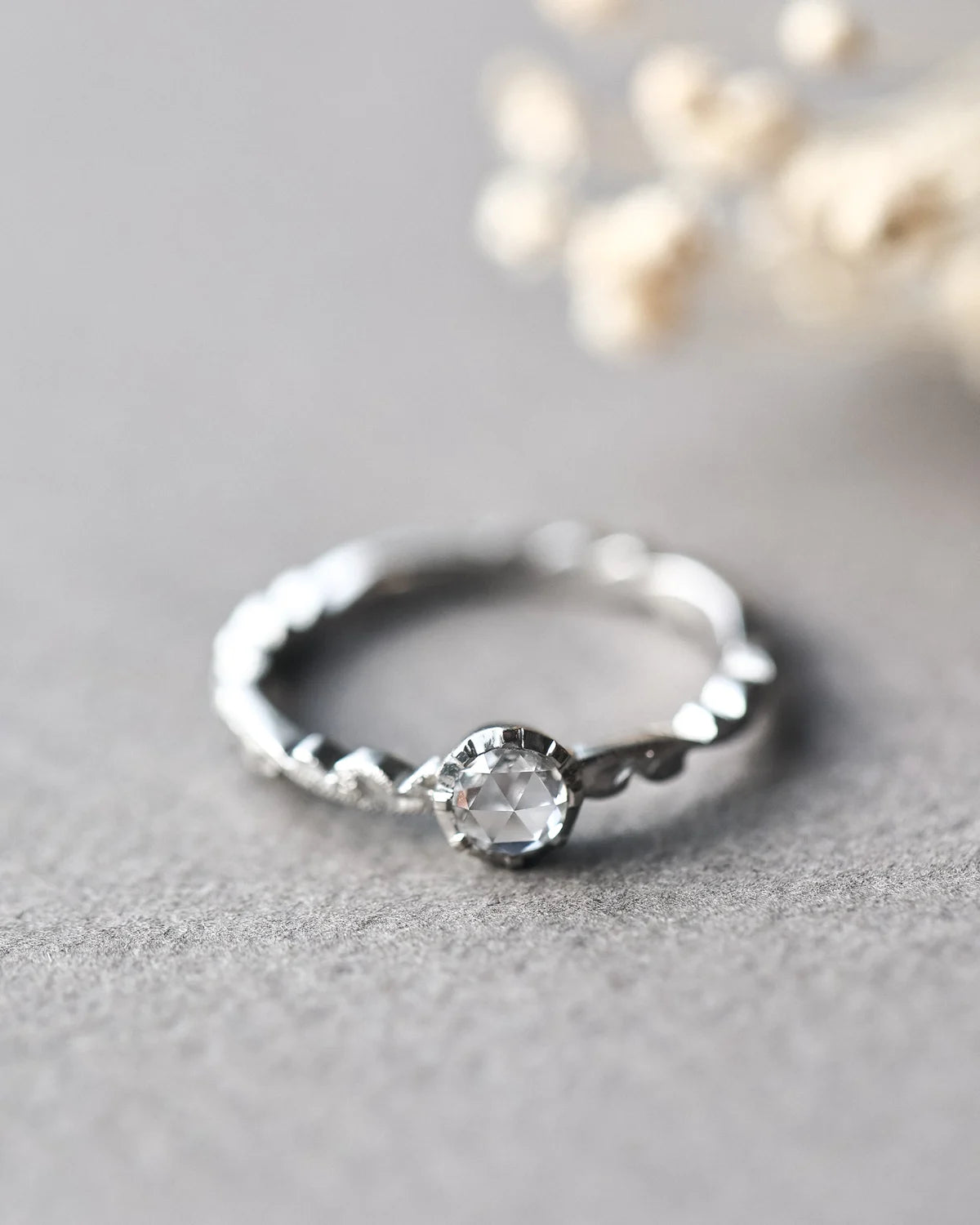 A silver ring with a textured, hammered band and a central clear faceted gemstone, resting on a textured gray surface with blurred dried flowers in the background.