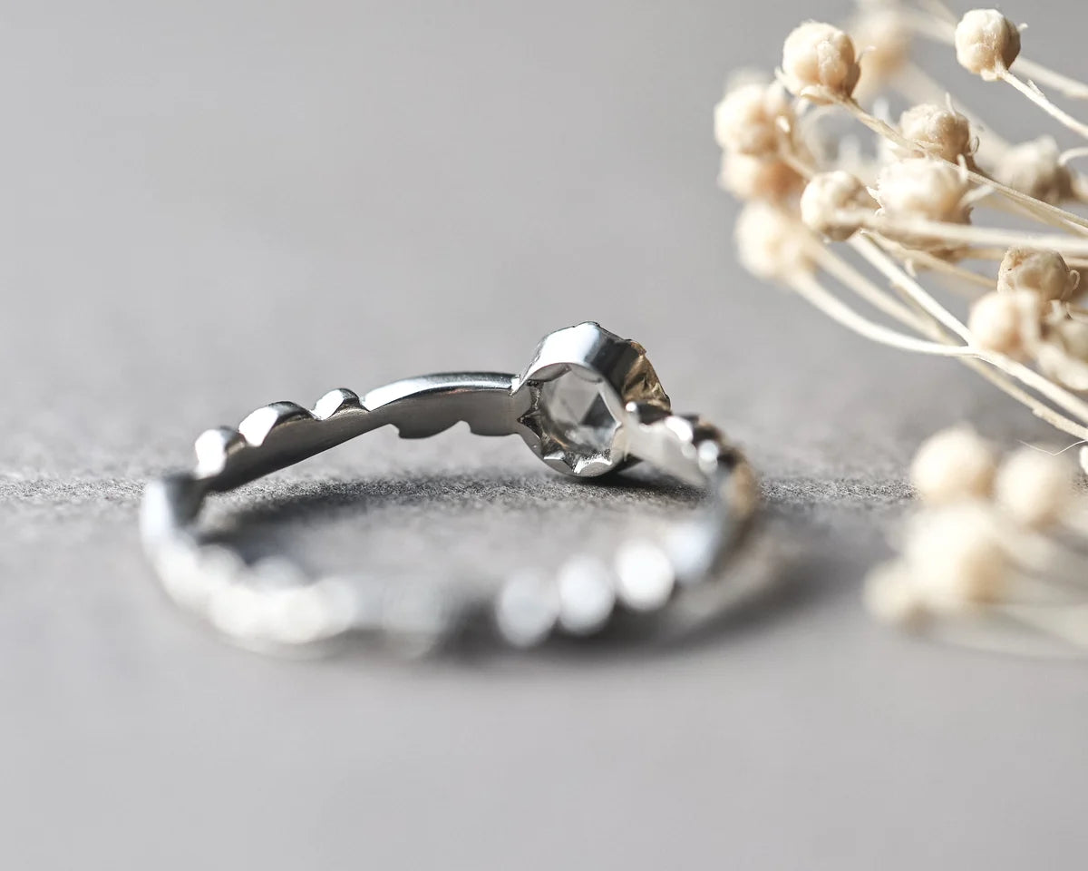 A silver ring with a textured, wavy band and a faceted dome-cut clear gemstone, resting on a gray surface with dried flowers in the background.