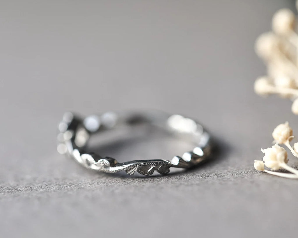 A silver twisted vine ring with engraved leaf details, resting on a textured gray surface with dried white flowers in the background.