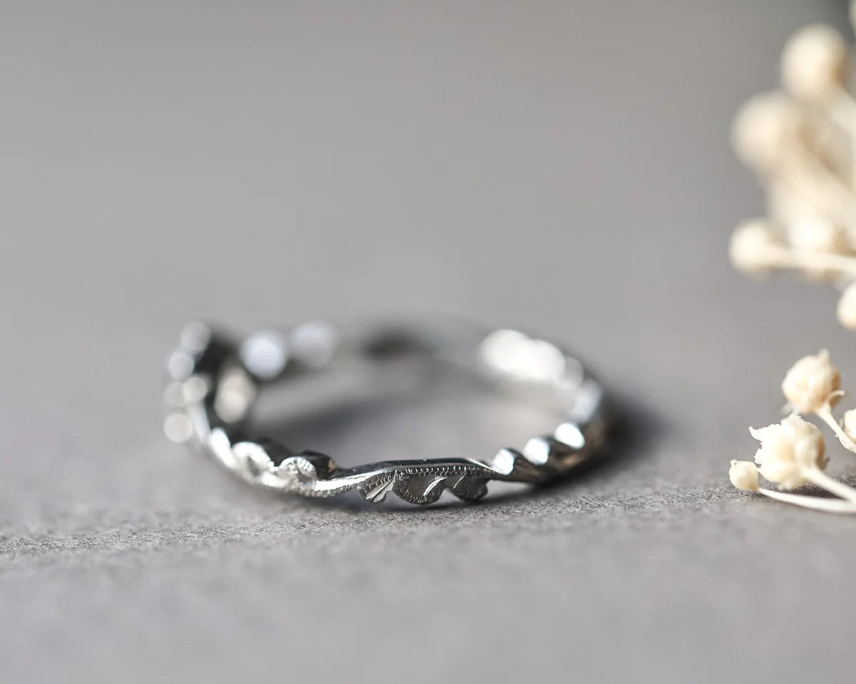 A silver twisted vine ring with engraved leaf details, resting on a textured gray surface with dried white flowers in the background.