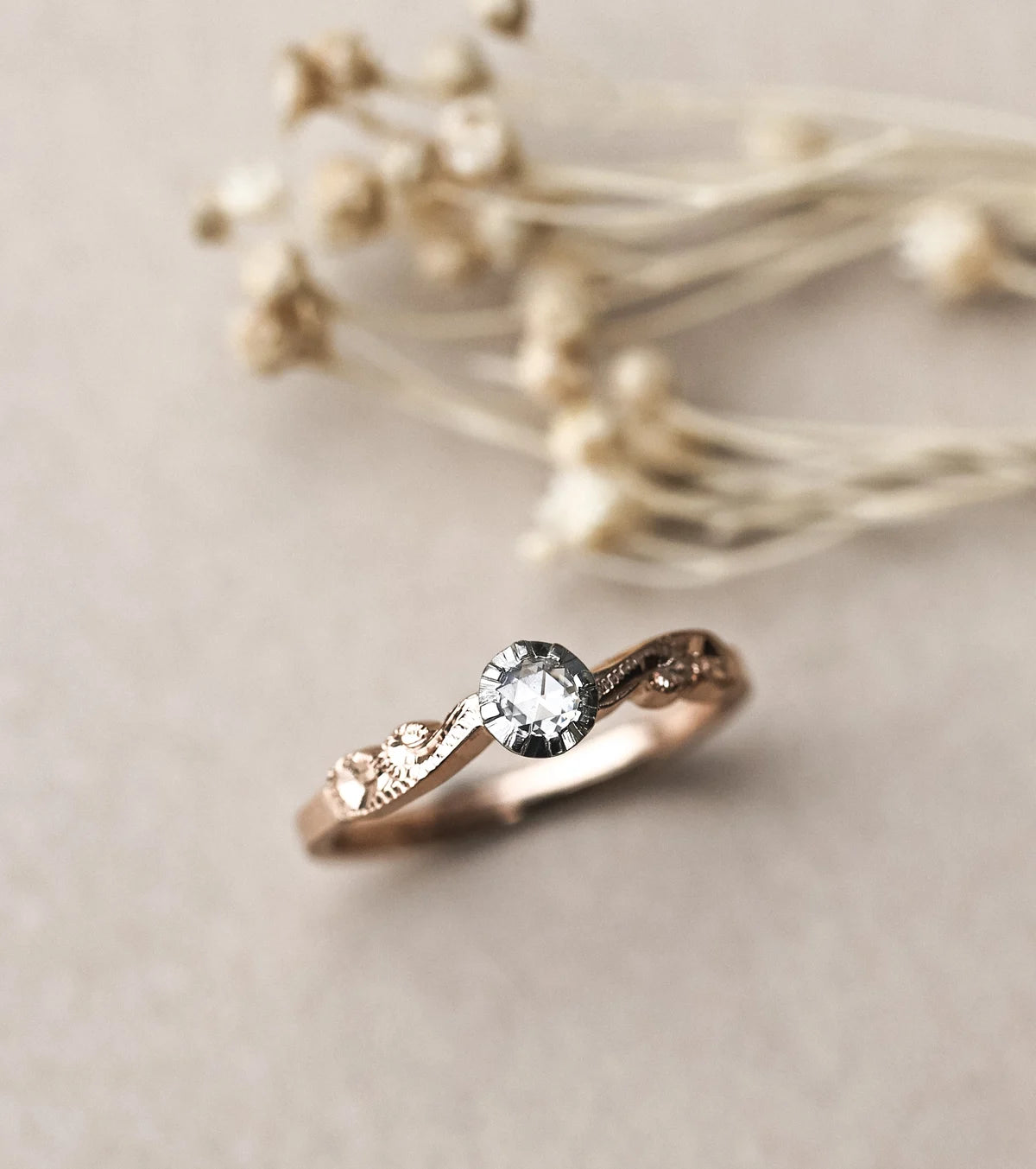 A rose gold ring with a central clear gemstone and small side stones, photographed on a soft beige surface with blurred dried flowers in the background.
