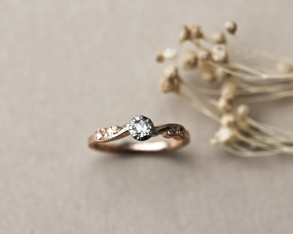 A rose gold engagement ring with a central round-cut clear gemstone and small side stones, styled on a neutral beige background with dried flowers.